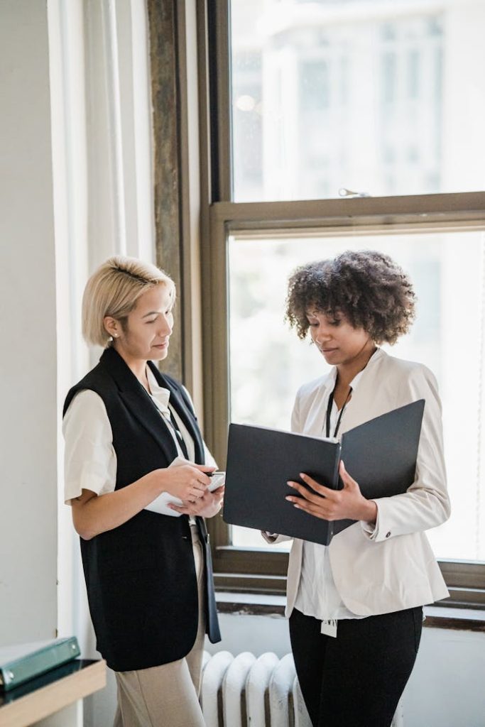 Mastering the First Impression: Your intriguing post title goes here Two women in business attire discussing documents by a window in the office.